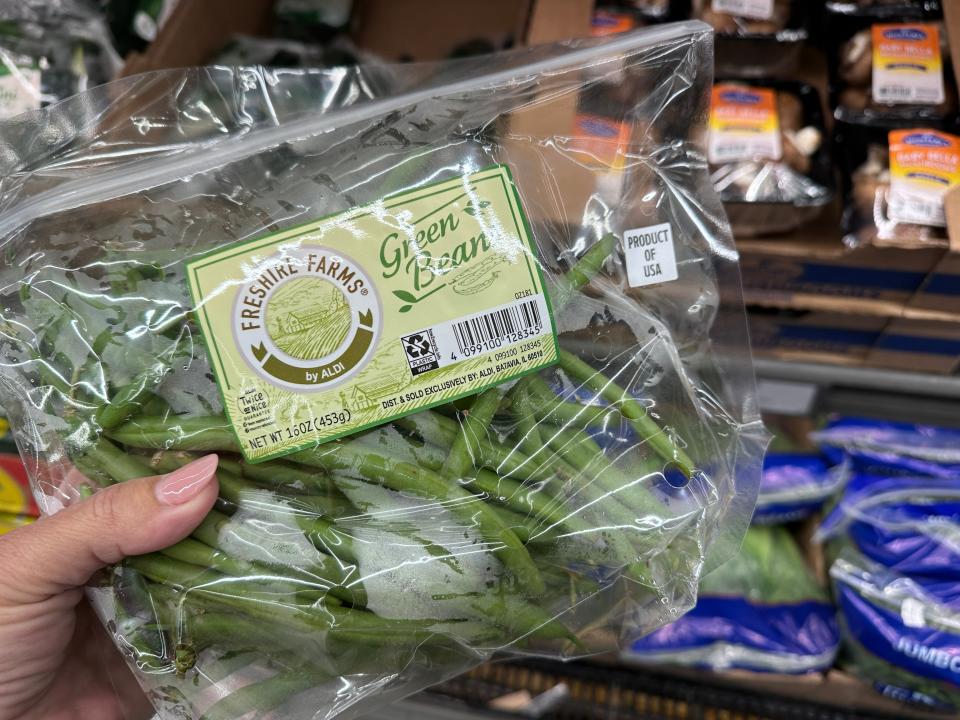 The writer holds a clear plastic bag of green beans in front of a shelf of produce at Aldi