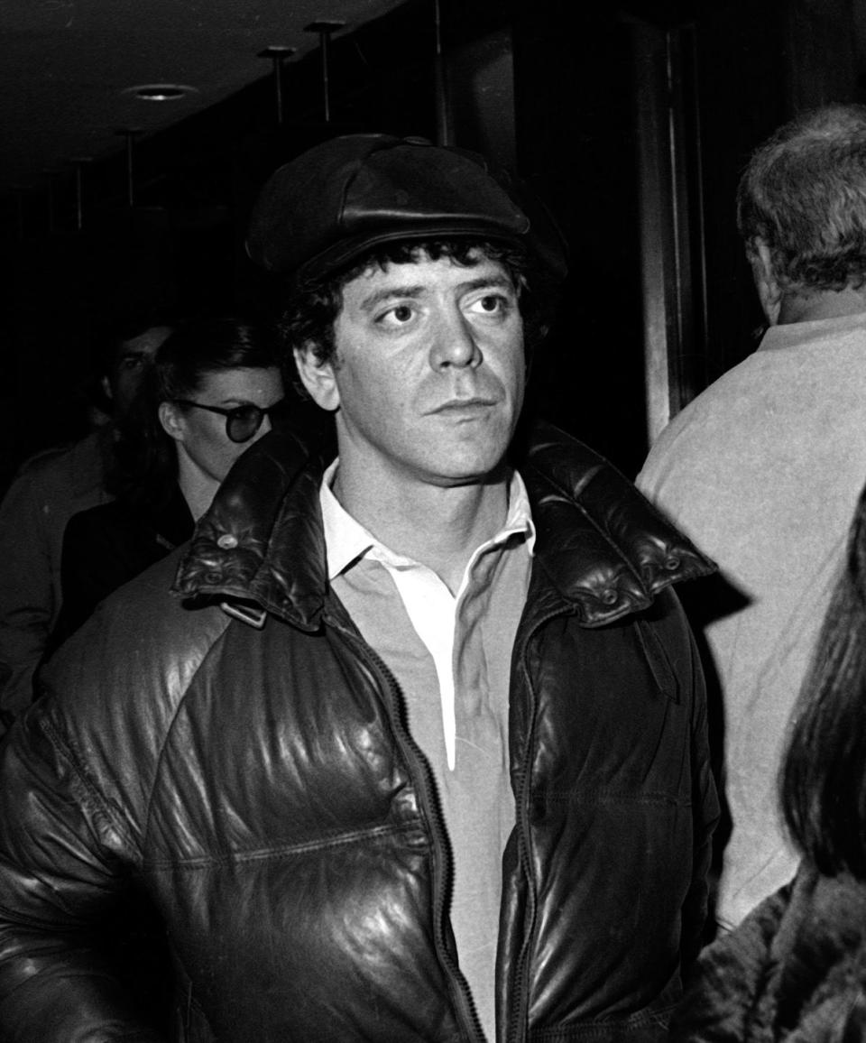lou reed stands near a few other people, he wears a puffy coat, hat, and collared shirt