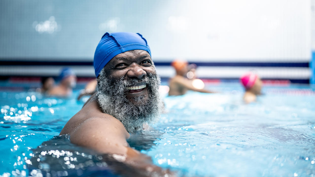  Older man with greyin beard wears a swim cap and smiles as he's swimming at a public pool. 