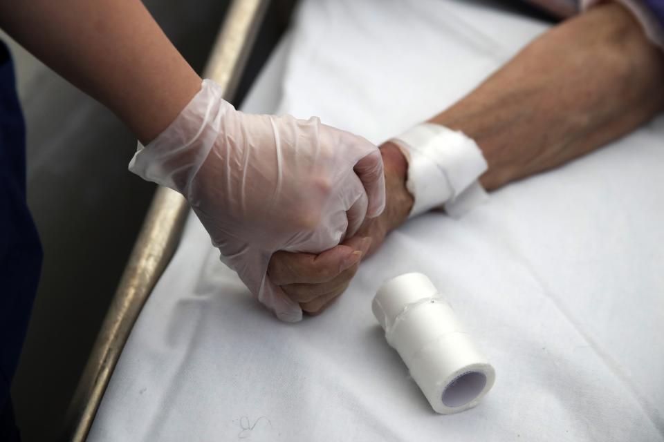 In this photo taken on Thursday, May 7, 2020, medical student Michaella Alexandrou holds the hand of a patient as her colleague takes a blood test at the Pathological Clinic of Sotiria Hospital in Athens. Greece's main hospital for the treatment of COVID-19 is also the focus of a hands-on training program for dozens of medical students who volunteered to relieve hard-pressed doctors from their simpler duties while gaining a close peek at the front lines of a struggle unmatched in modern medical history. (AP Photo/Thanassis Stavrakis)