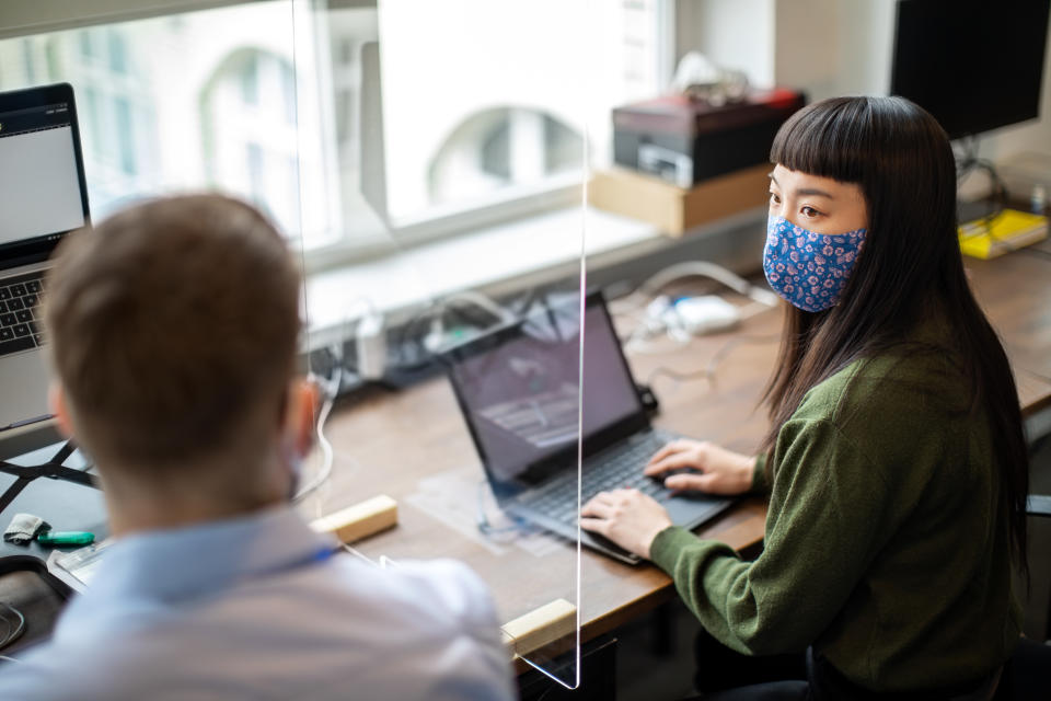 Colleagues discussing work through glass partition at office desk. Businessman and businesswoman sitting at desk with protection guard between them discussing work post covid-19 pandemic.