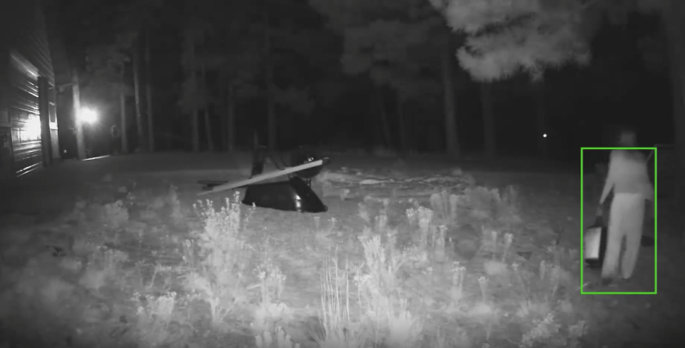 A person in night vision security footage carries an object while walking near a wooded area.  A green box highlights the person