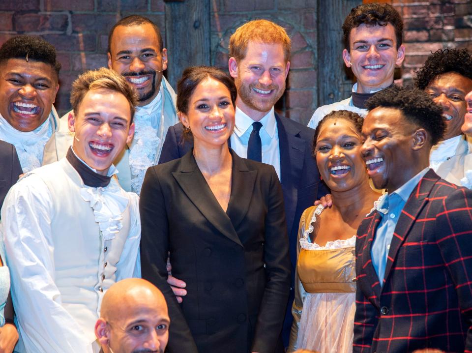 Meghan standing in front of Harry and both are smiling while the "Hamilton" cast smile and laugh around them.