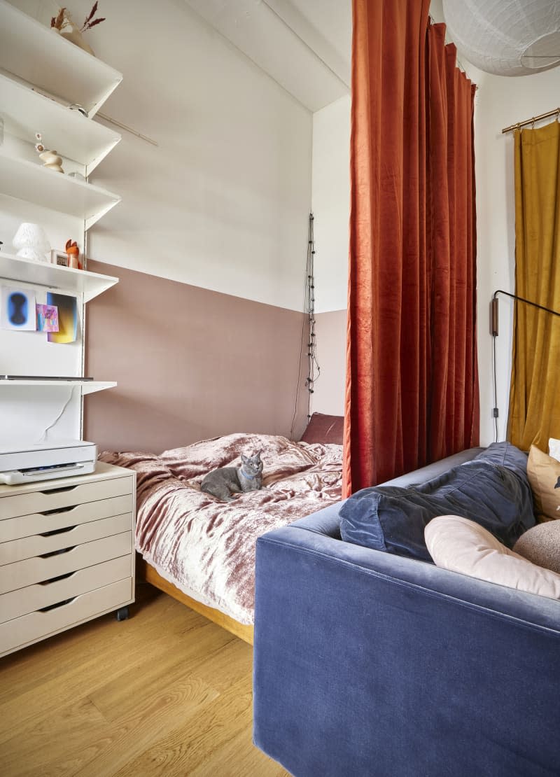 bedroom area separated from living area by orange curtain, muted rose colored wall, white shelves, blue velvet sofa