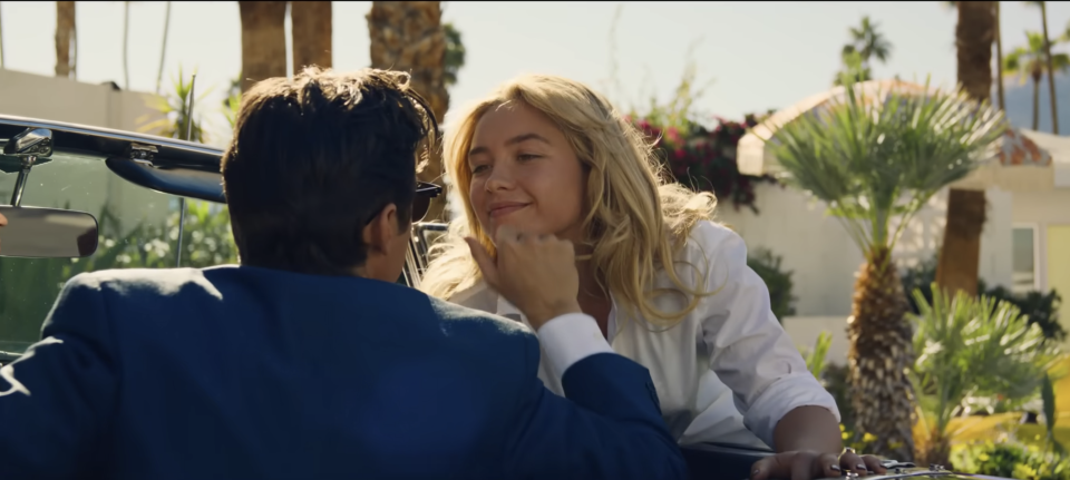 A man in a convertible, his back to us, caresses the face of a woman standing over the car and smiling