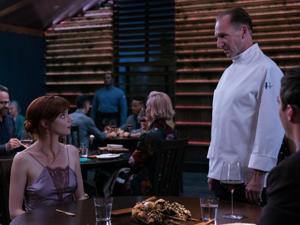 Anya Taylor-Joy and Ralph Fiennes in The Menu with one seated and the other standingin a restaurant