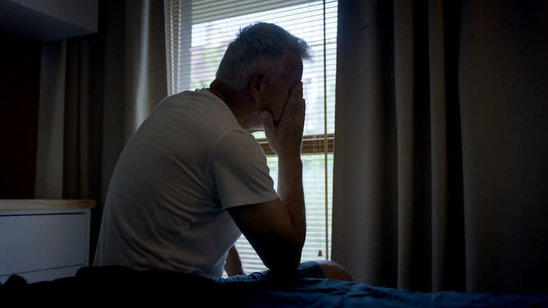 Man in a white t-shirt sitting on a bed, hands covering his face, looking distressed while facing a window with closed blinds