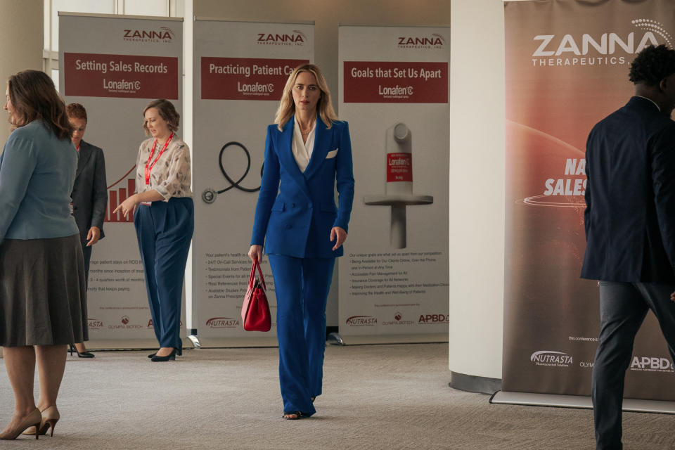 Emily Blunt in Painkiller walking past posters for Zanna, the pharmaceutical company