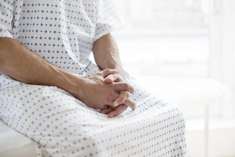 A person sits with clasped hands, wearing a hospital gown with a simple pattern, in a medical setting