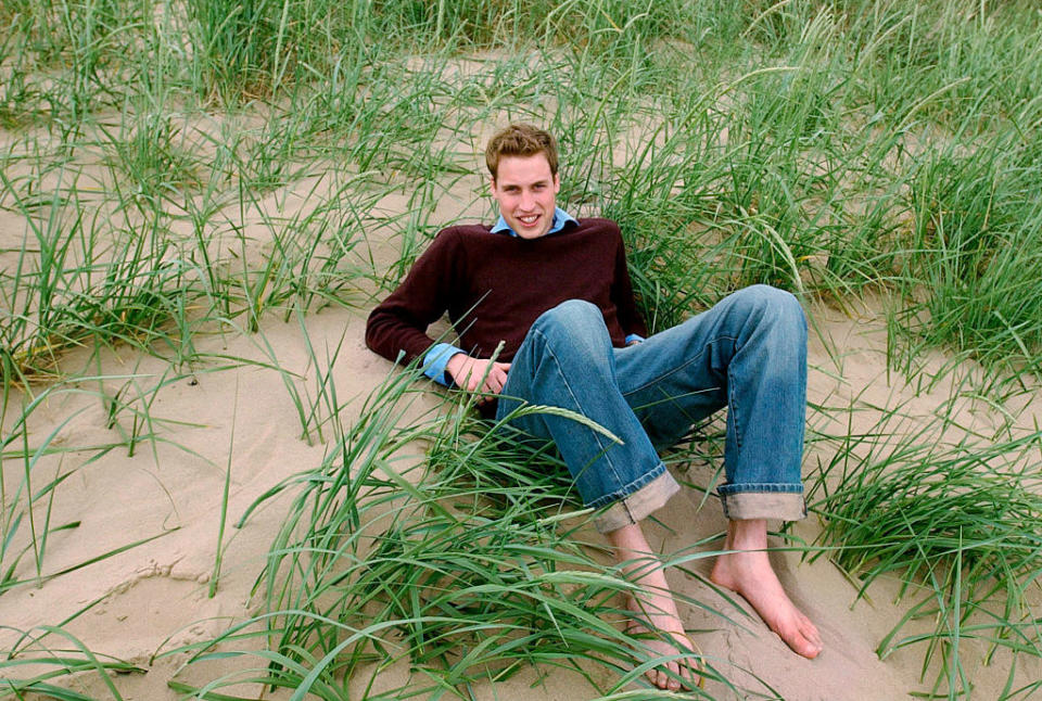He's smiling and leaning back on the grassy sand in jeans and a sweater and barefoot, with his knees bent