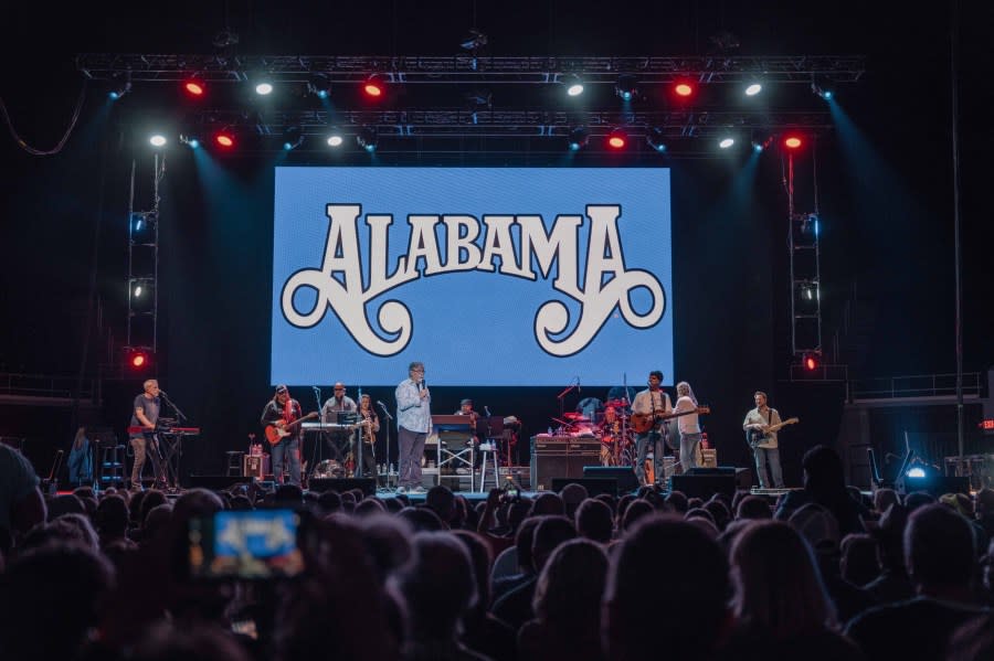 Photo of a concert. There is an Alabama music group logo on a blue screen. On the stage you can see the group standing with microphones and various musical instruments.