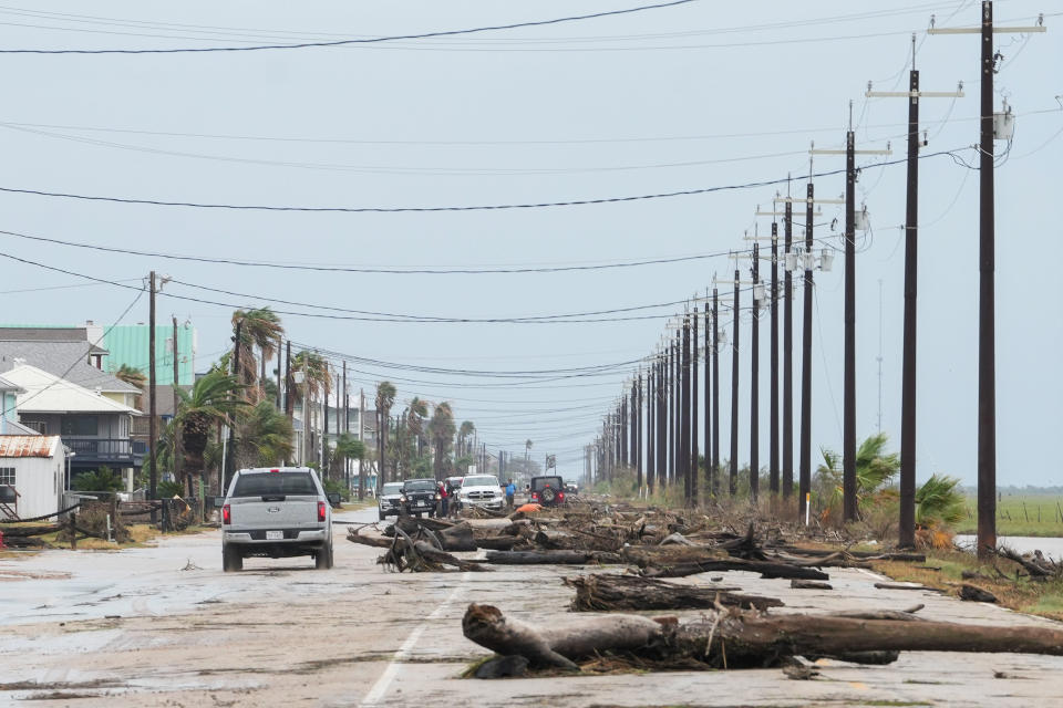 A road damaged by a storm with downed trees and power lines, cars trying to navigate around debris, and houses in the background