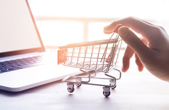 A person's hand pushes a miniature grocery cart in front of an open laptop computer.