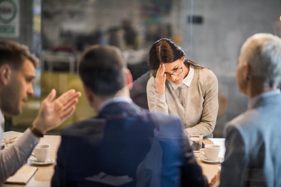 Four people are sitting around a desk in what appears to be a meeting. One person, looking stressed or overwhelmed, touches their forehead with a hand. Cups of coffee are on the table