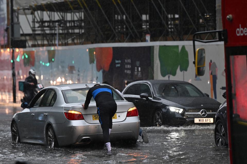 A person pushes a broken-down car through deep water on a flooded road in The Nine Elms district of London on July 25, 2021 during heavy rain. - Buses and cars were left stranded when roads across London flooded on Sunday, as repeated thunderstorms battered the British capital. (Photo by JUSTIN TALLIS / AFP) (Photo by JUSTIN TALLIS/AFP via Getty Images)