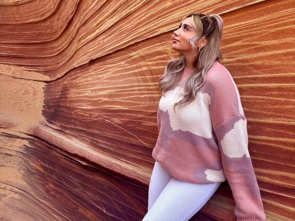 The writer wears white pants and a pink and white sweater and leans against rock wall with orange and red layers and looks up