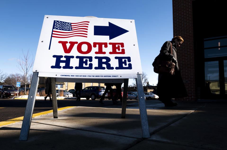 A person, looking down, stands on a sidewalk near parked cars and a sign that reads: Vote here