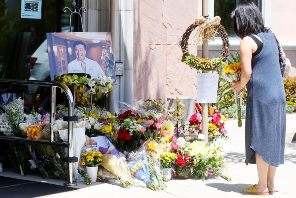 A woman stands before a flower-filled memorial outside a building. Amid the flowers, is an easel carrying the portrait of a man.