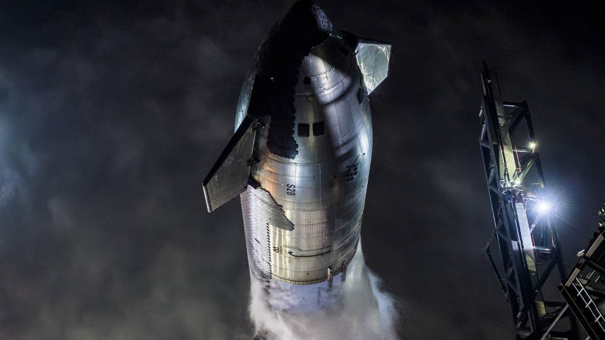  A large black and silver rocket vents vapor at night while standing upright on a launch pad. 