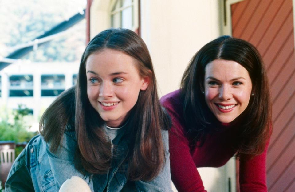 Rory and Lorelai from "Gilmore Girls" lean forward, smiling, in casual attire