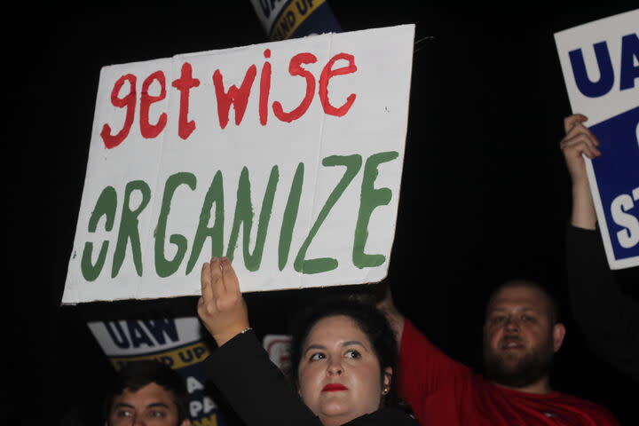 A woman holds up a sign saying "Get wise organize" on a picket line. "Get wise" is written in red; "Organize" is written in green.