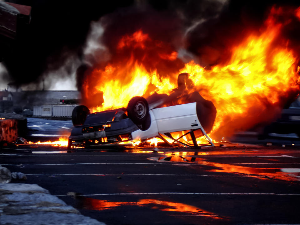 A white car is overturned and engulfed in flames on a street at night, with large clouds of smoke rising into the sky