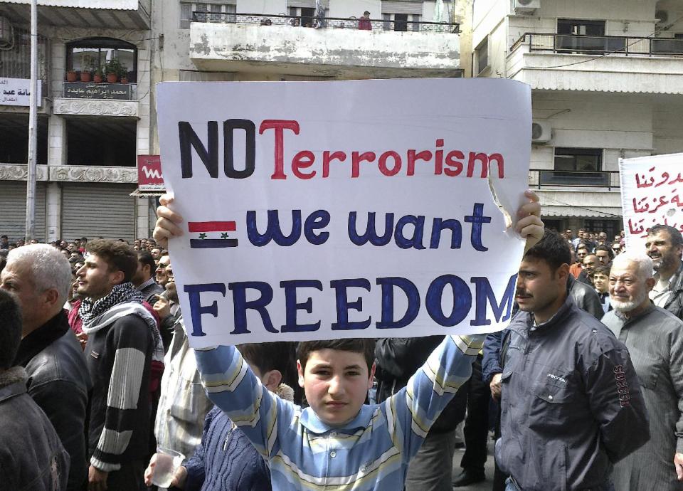 FILE - In this April 22, 2011, file citizen journalism image made on a mobile phone and acquired by the AP, a Syrian boy carries a banner during an anti-government demonstration in the coastal city of Banias, Syria. The sectarian divisions burning across the rest of Syria have become physical barriers in this town on the Mediterranean coast, with soldiers manning sandbagged checkpoints dividing its Sunni Muslim southern neighborhoods from those of the Alawite and Christians in the north. Yet even after a mass killing of Sunnis in a village outside Banias in May, a tenuous truce keeps this town of 50,000 people locked in a frigid, divided peace in hopes the chaos engulfing the rest of the country will pass them by.(AP Photo, File)