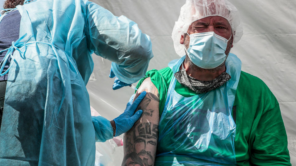 Joseph Kuresa, wearing a face mask and hair covering, winces as he receives a shot in his upper arm. 