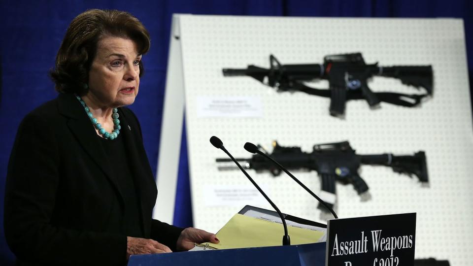 dianne feinstein speaking into a microphone at a podium, with assault weapons mounted on a display behind her