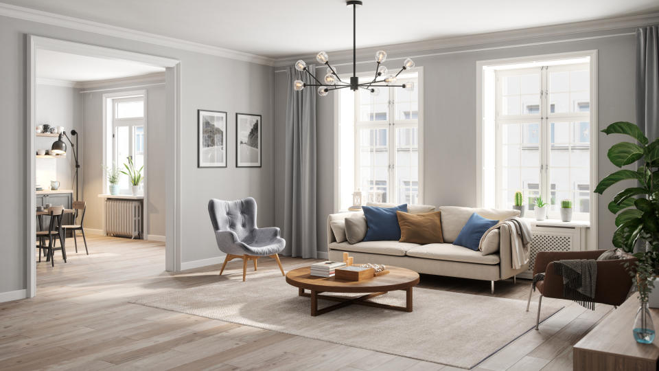 Modern living room with large windows, a beige sofa, a gray armchair, a wooden coffee table, and wall decor. Dining area and home office visible in background