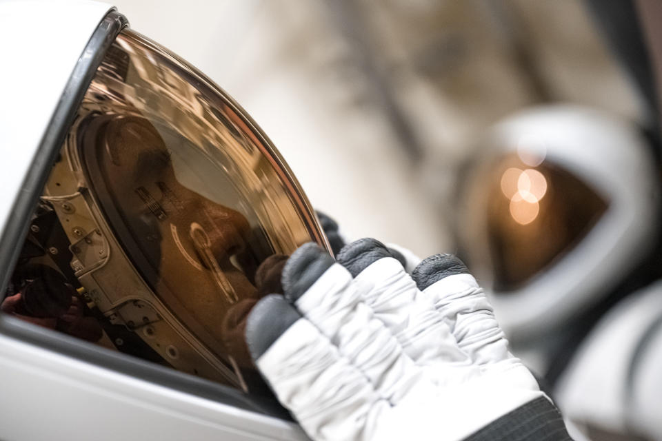 A close-up of an astronaut helmet with a white shell extending from left to center of image. A copper-shaded visor runs along the pale interior surface. A white glove with a black tip tests the visor.