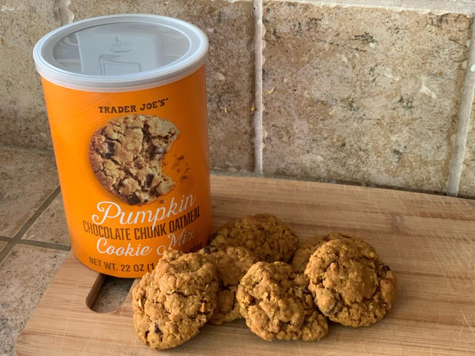 Orange container of Trader Joe's pumpkin spice oatmeal chocolate chip cookie mix beside baked cookies on a wooden cutting board