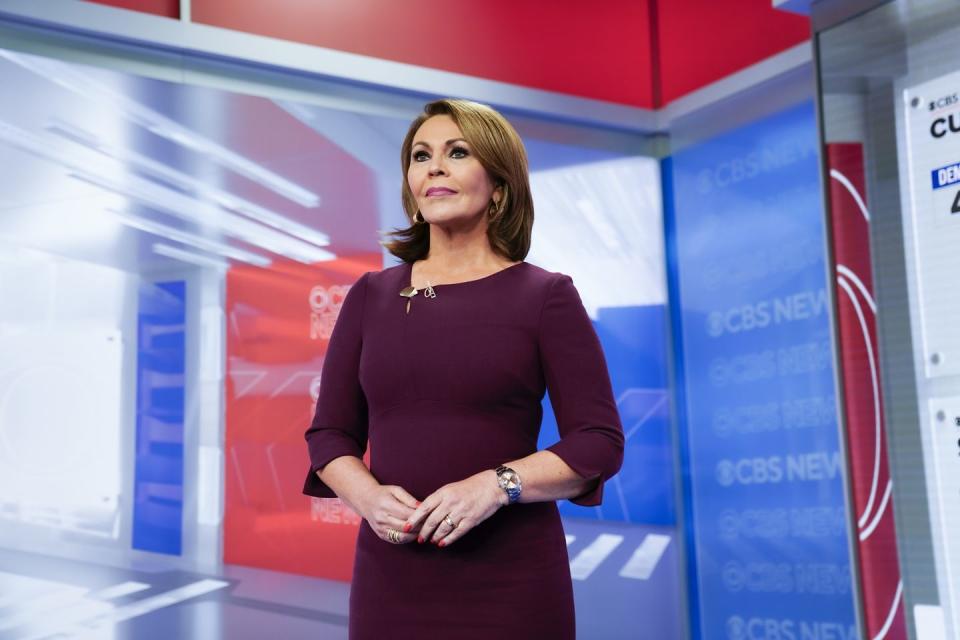 maria elena salinas stands on a cbs news television set with red, white, and blue backgrounds, she wears a purple long sleeve dress with a watch and hoop earrings