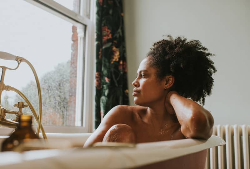 A beautiful woman bathes in a roll top bath. She seems distracted as she rests her head on her arm and gazes out the window. Space for copy.