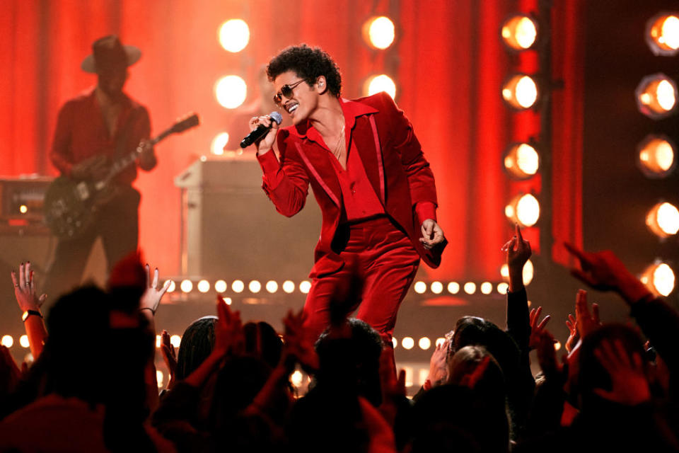 Bruno Mars performs on stage in a red suit, holding a microphone, with a guitar player in the background and an audience raising their hands