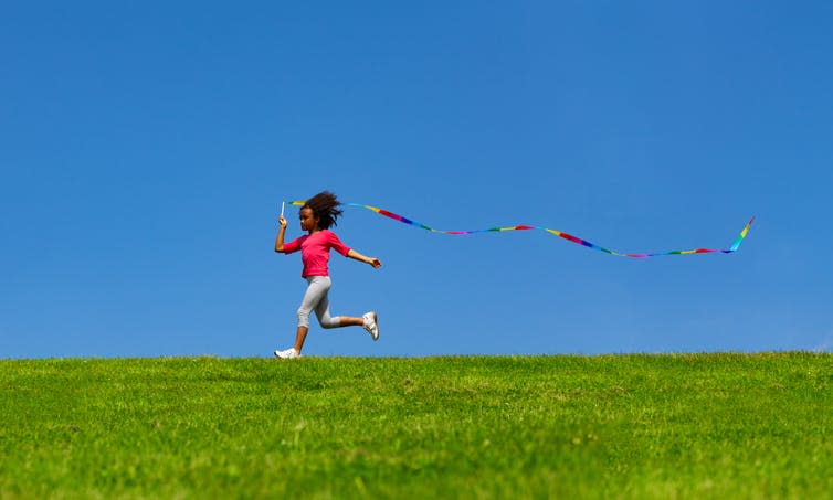 Young girl running on grass with a ribbon flowing out behind her against a bright blue sky.