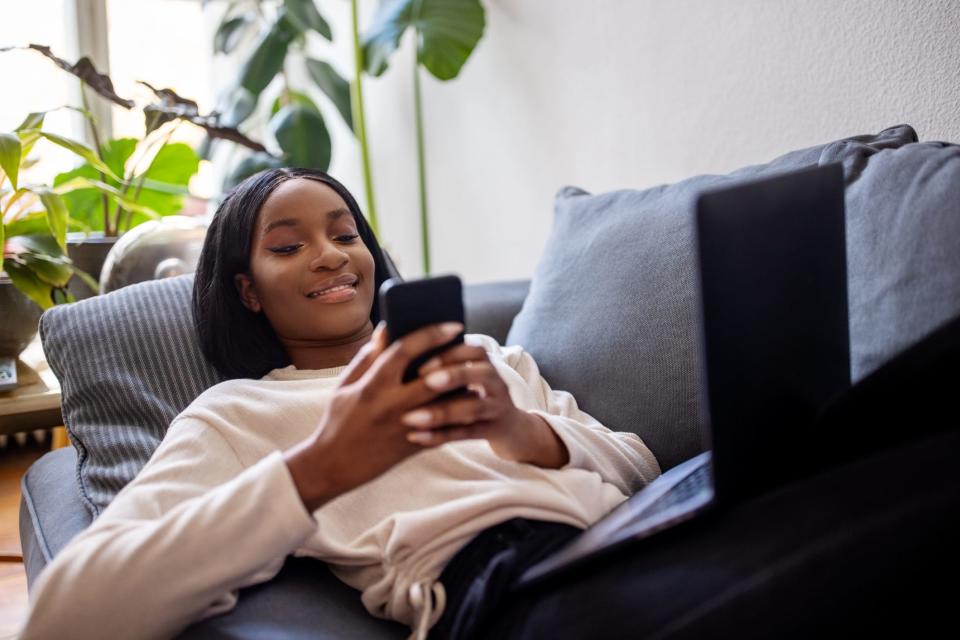 A woman is reclining on a couch and smiling while looking at her phone.