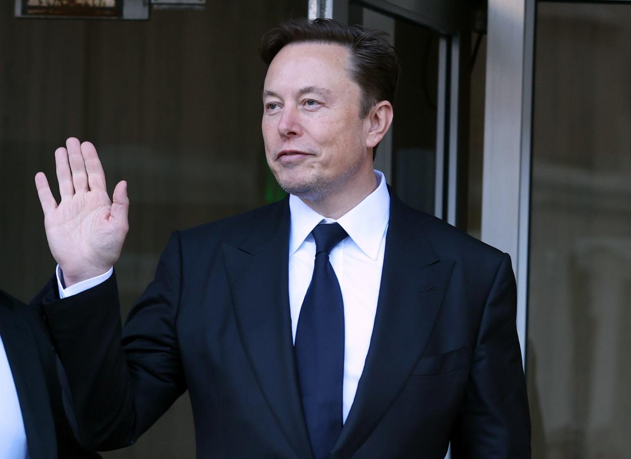 Elon Musk, wearing a black suit and tie, waves to photographers outside a courthouse