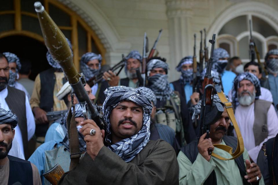 A dozen or more men with large weapons stand in front of a home with arched windows