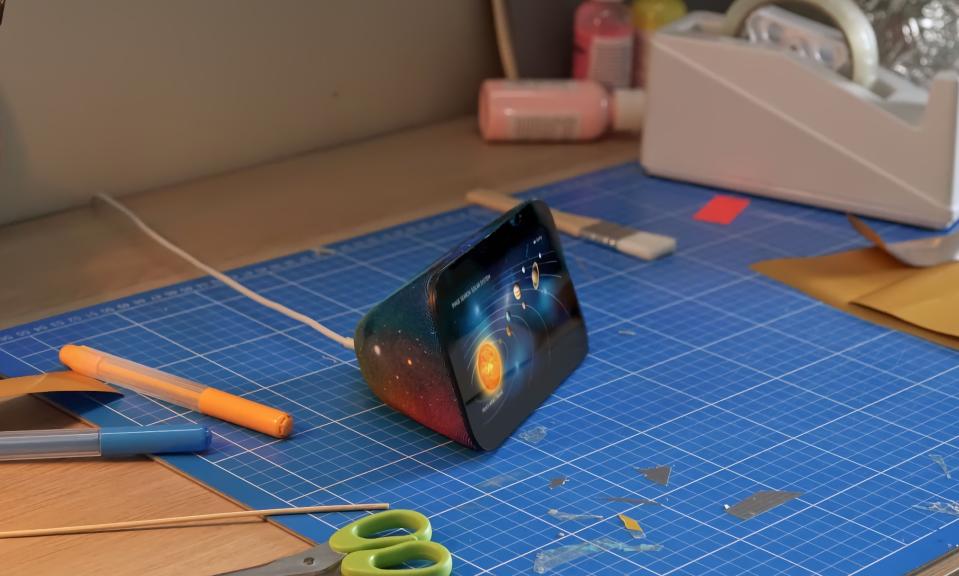 Amazon marketing photo of the Amazon Echo Show 5 Kids with a galaxy design on its back and an illustration of the solar system on its screen. It sits on a blue drafting mat on a desk with scissors, pens and other supplies nearby.