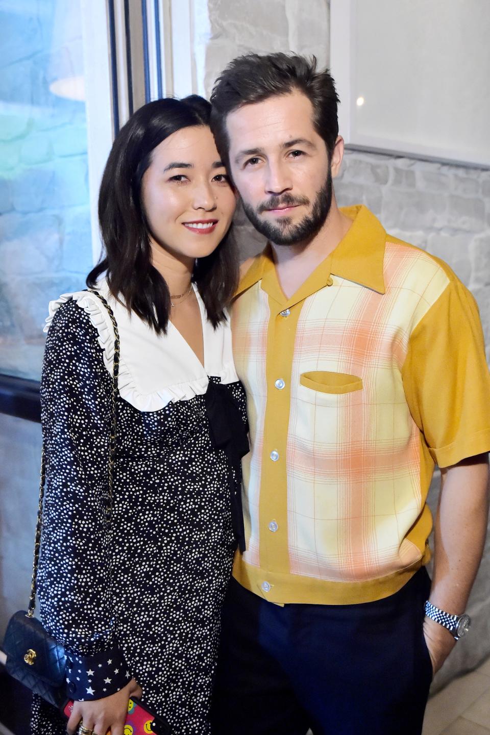 Maya Erskine, in a printed dress with a white collar, and Michael Angarano, in a vintage-style plaid shirt, pose together at an event