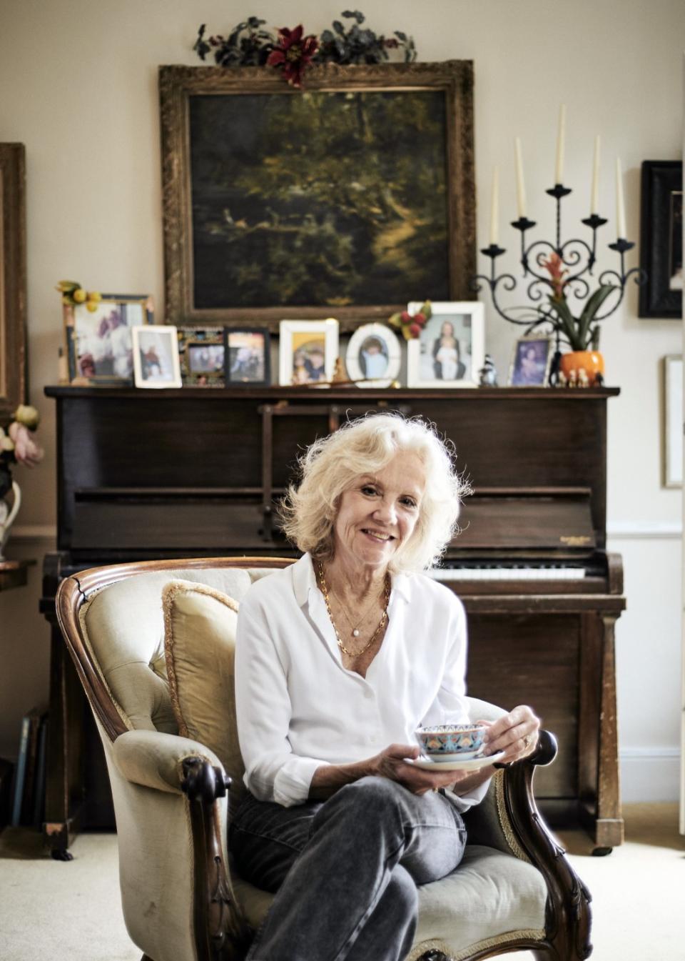 A woman sits on a living room chair in front of a piano with photos on top of it