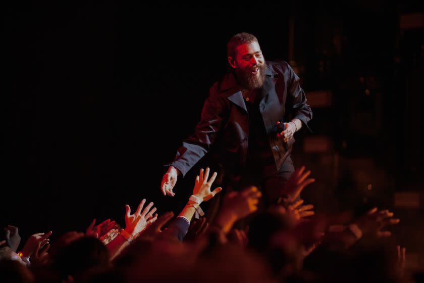 Post Malone, bearded, wearing black jacket reaching out to fans from the stage with smile on his face