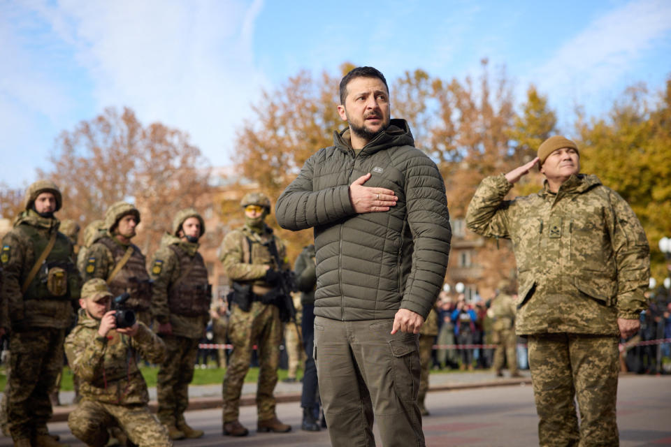 Zelensky, in cargo pants and a hooded down jacket, stands with his right hand over his heart among half a dozen soldiers in helmets and camouflage fatigues, one of whom trains a camera at Zelensky.