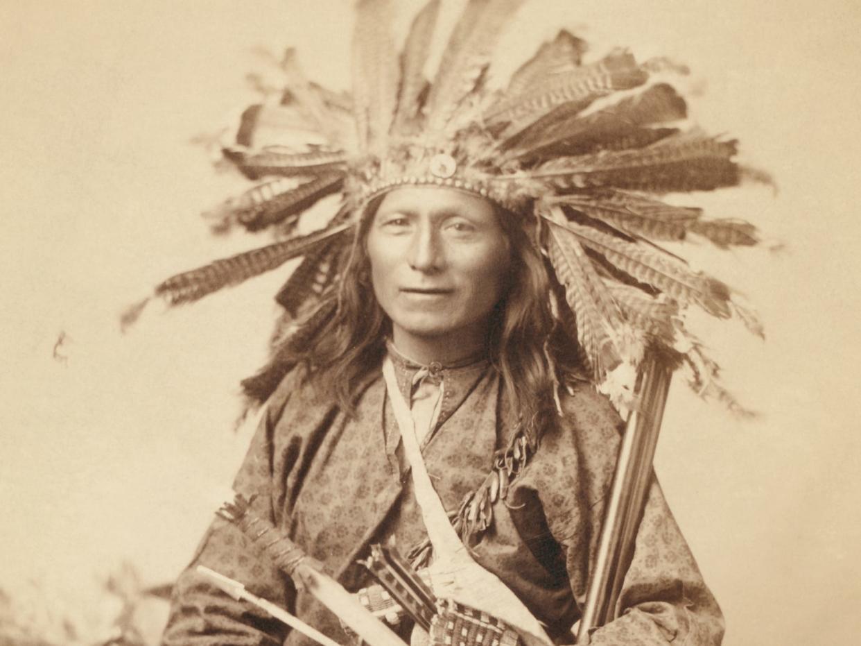 Little, Oglala band leader, three-quarter length studio portrait, seated, wearing a turkey feather headdress and holding various weapons