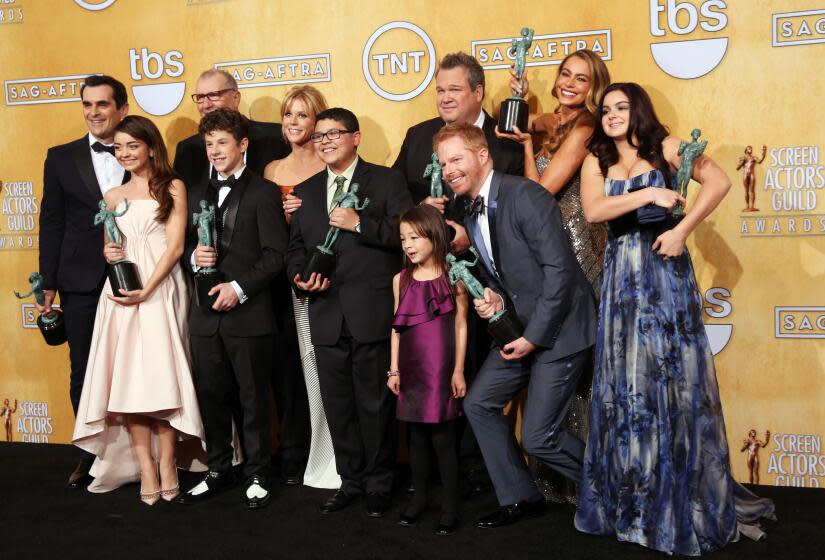 Cast of Modern Family, dressed in suits and dresses, huddle and pose together while holding SAG awards