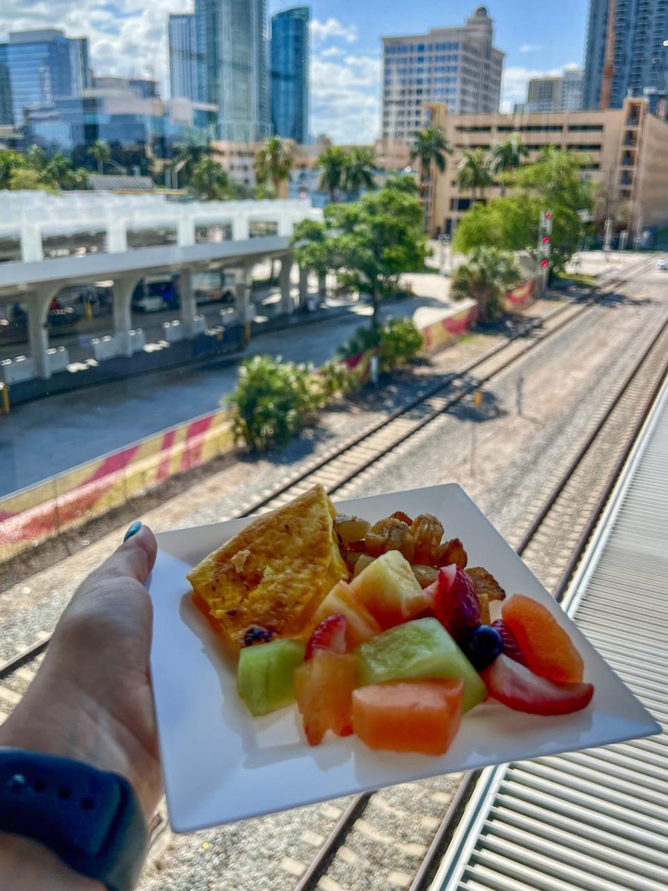 A hand holds a small plate with breakfast food is held up in front of a window overlooking the Brightline train tracks.