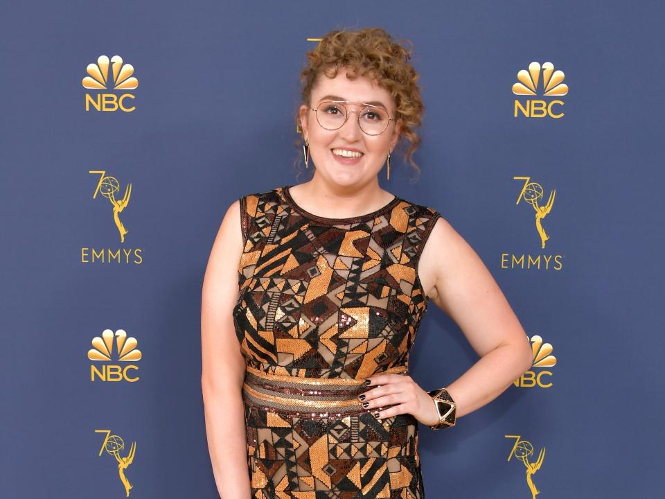 comedian emily heller in a geometrically patterned dress on the 2018 emmys red carpet, smiling with one hand on her waist