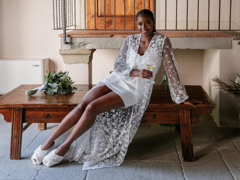 A bride sits on a bench in a robe. Her bouquet sits next to her.