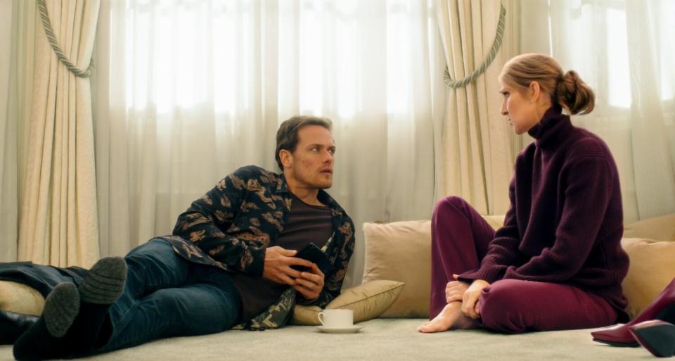 Sam Heughan and Celine Dion sit on a couch in front of a curtained window.